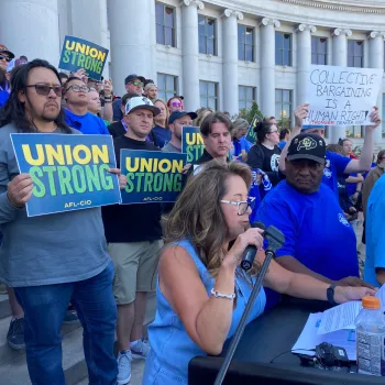 A woman holding a microphone in front of a crowd holding "union strong" signs